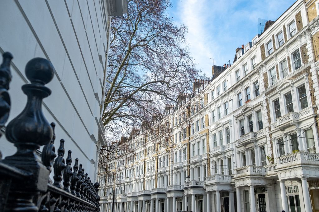 View of a city street with white multi-story townhouses lined with balconies, seen from behind a black iron fence; a leafless tree and blue sky in the background.